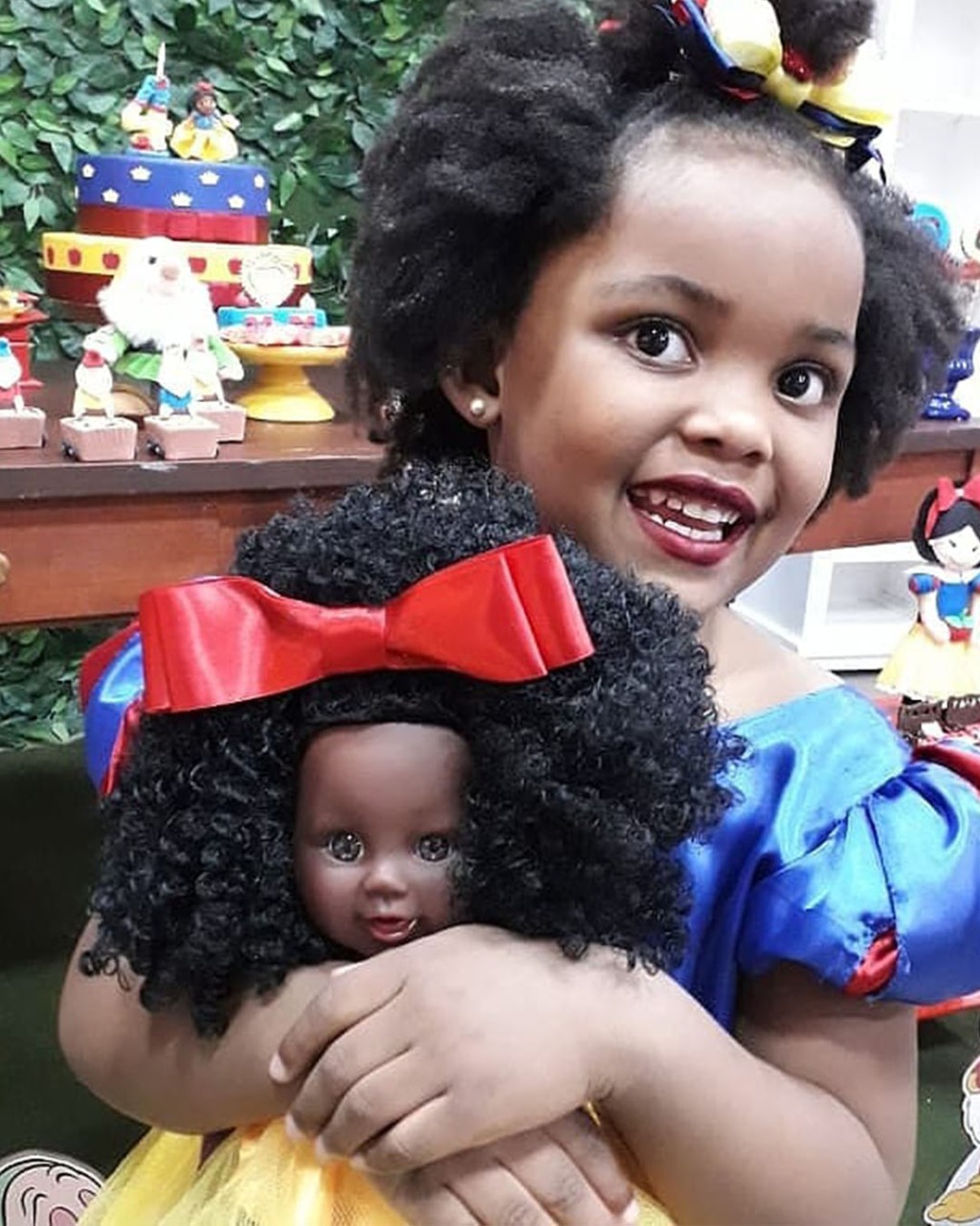 Menina vestida de branca de neve sorrindo enquanto segura uma boneca negra também usando fantasia de branca de neve