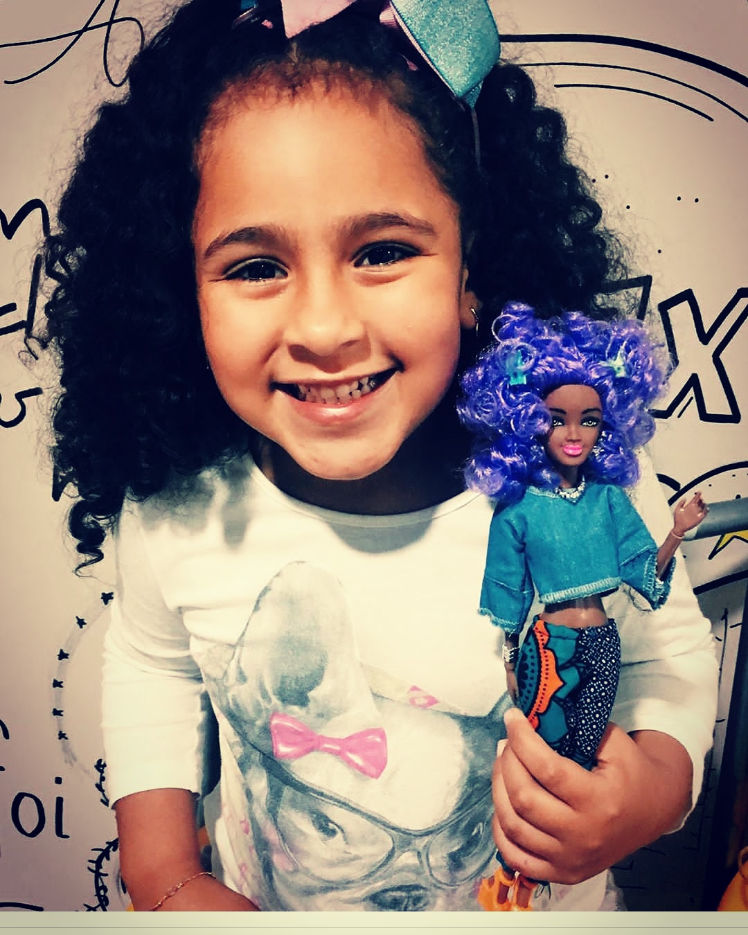 Menina de camiseta branca sorrindo enquanto segura uma boneca de cabelo azul e roupas azuis